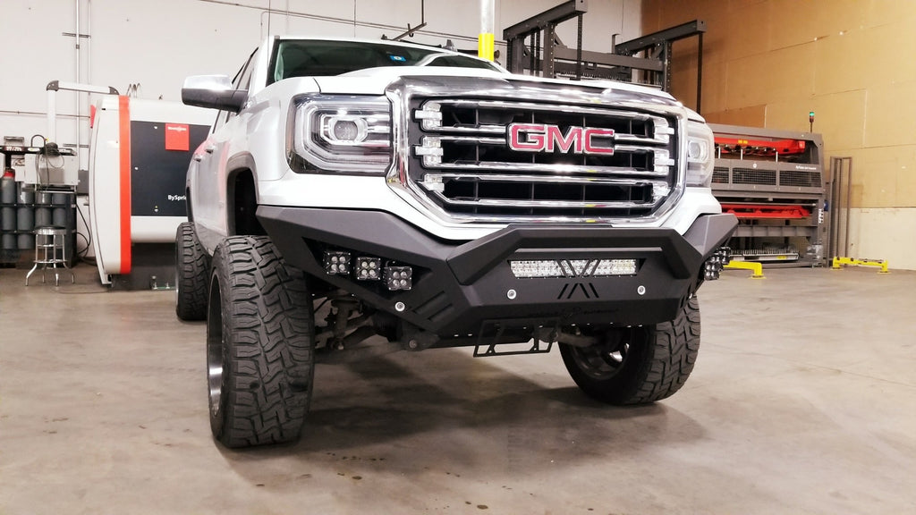 A white lifted GMC 1500 with oversized off-road tires and a 2016-2018 Spartan Front Bumper in textured black is parked inside a spacious industrial garage, equipment and tools visible in the background.