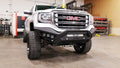 A white lifted GMC 1500 with oversized off-road tires and a 2016-2018 Spartan Front Bumper in textured black is parked inside a spacious industrial garage, equipment and tools visible in the background.