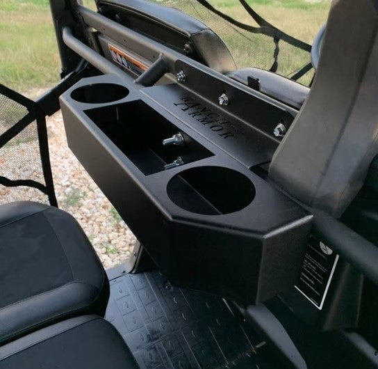 Close-up of a Can-Am Defender Rear Cup Holder Basket, labeled JAWKOR, installed between two vehicle seats. The black console features two cup holders and a shallow rectangular compartment. Mesh netting and grassy terrain are in the background.