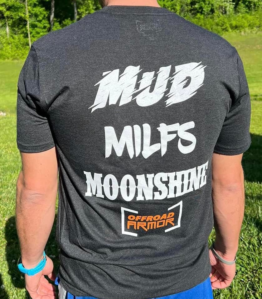 A person stands on grass, facing away, wearing an Offroad Armor T-Shirt featuring bold white text: MUD, MILFS, and MOONSHINE, plus an orange and black Offroad Armor logo at the bottom. Trees and sunlight appear in the background.