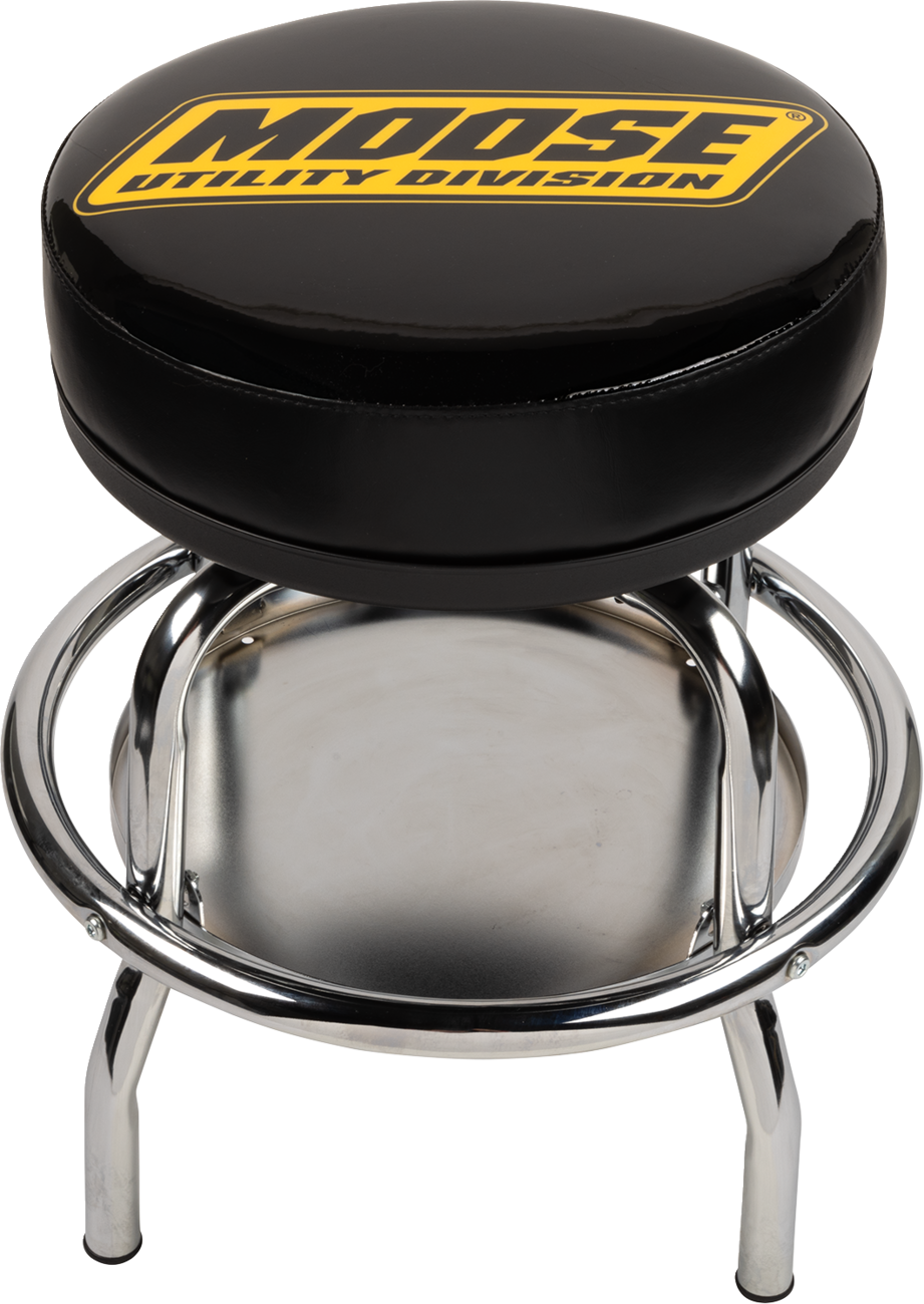 A black round swivel stool with chrome legs and a circular footrest. The cushioned seat features bold yellow text reading “MOOSE UTILITY DIVISION.” There is a metal tray below the seat between the stools legs.