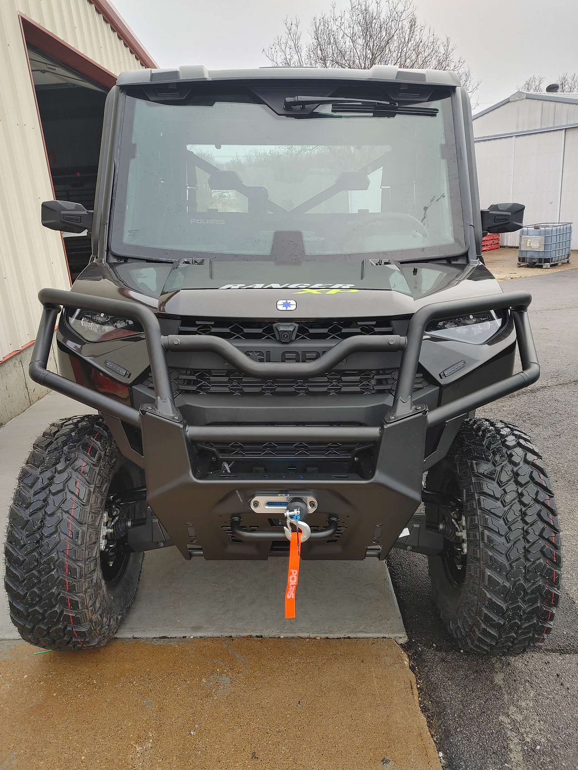 A Polaris Ranger 1000 XP (2018-Present) equipped with a Power Harness Turn Signal Kit is parked on concrete outside a metal building, featuring rugged tires, a sturdy front bumper with winch and orange tow strap, and an oversized windshield.