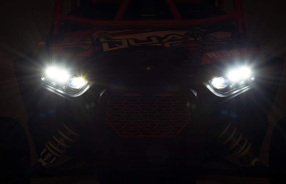 A close-up view of an off-road vehicle with its bright headlights turned on, illuminating the dark surroundings. The image shows the vehicles front grill, suspension springs, and partial red accents on the frame.