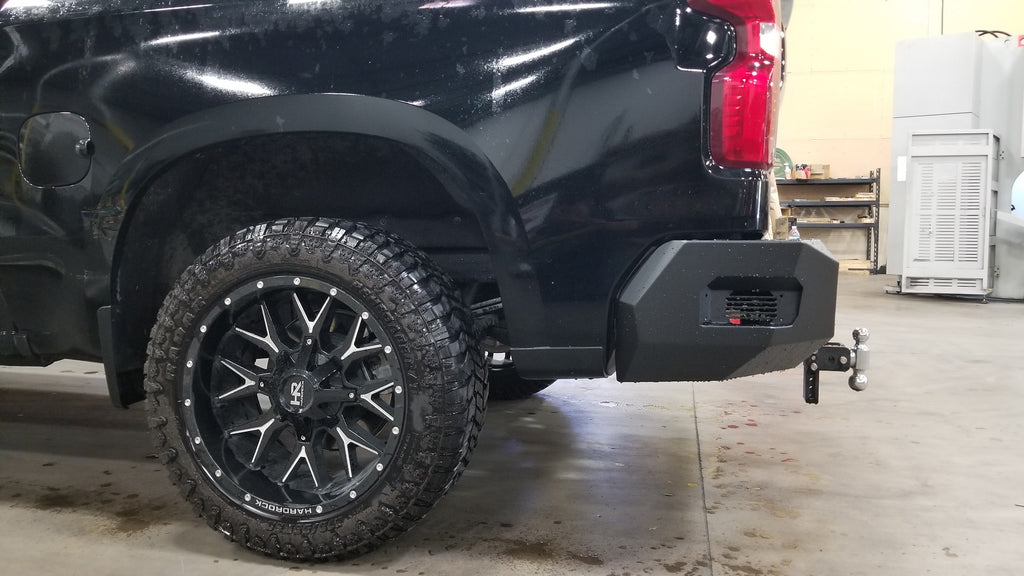 A close-up side view of a black pickup shows the 2019-2021 Chevy 1500 Spartan Rear Bumper in texture black, rugged off-road tire with black alloy rim, and trailer hitch indoors on concrete with workshop equipment in the background.