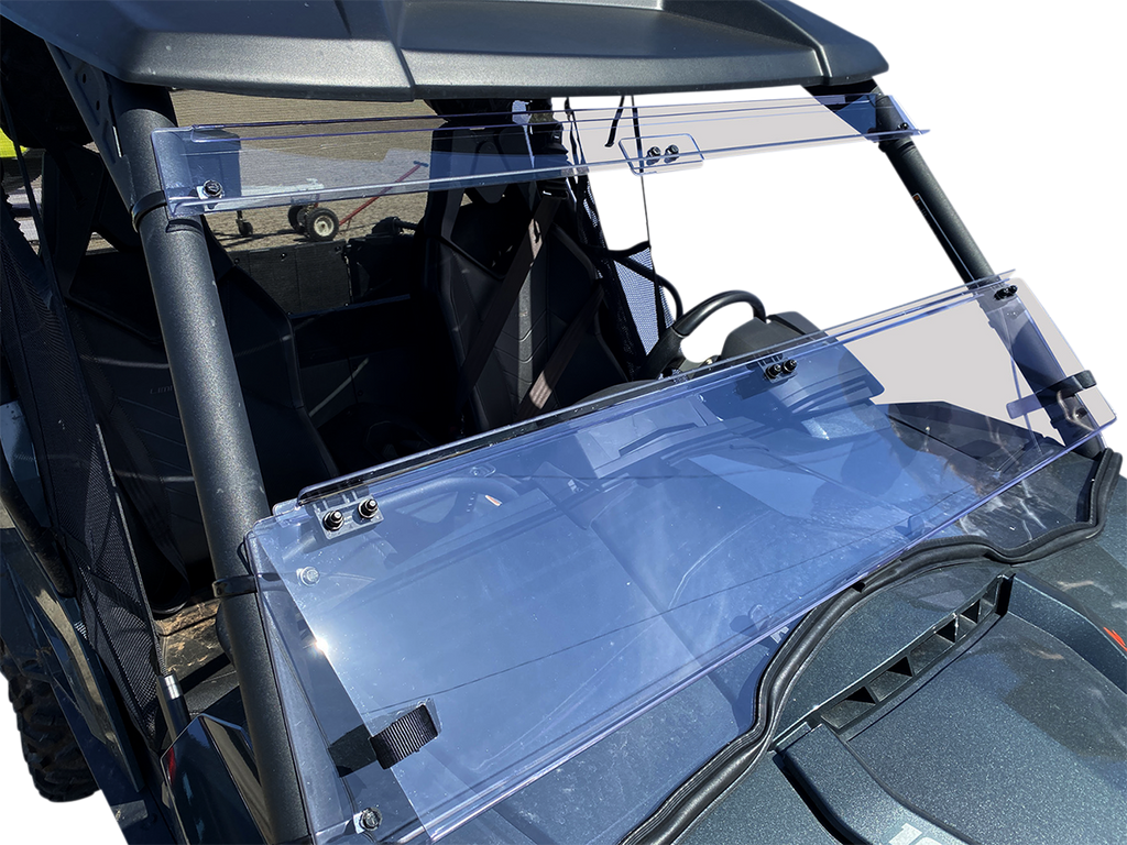 A close-up view of a utility vehicle’s clear polycarbonate folding windshield, mounted on a black roll cage. The windshield is partially open, showing two hinge sections and metal mounting brackets. The seats and steering wheel are visible behind the wind