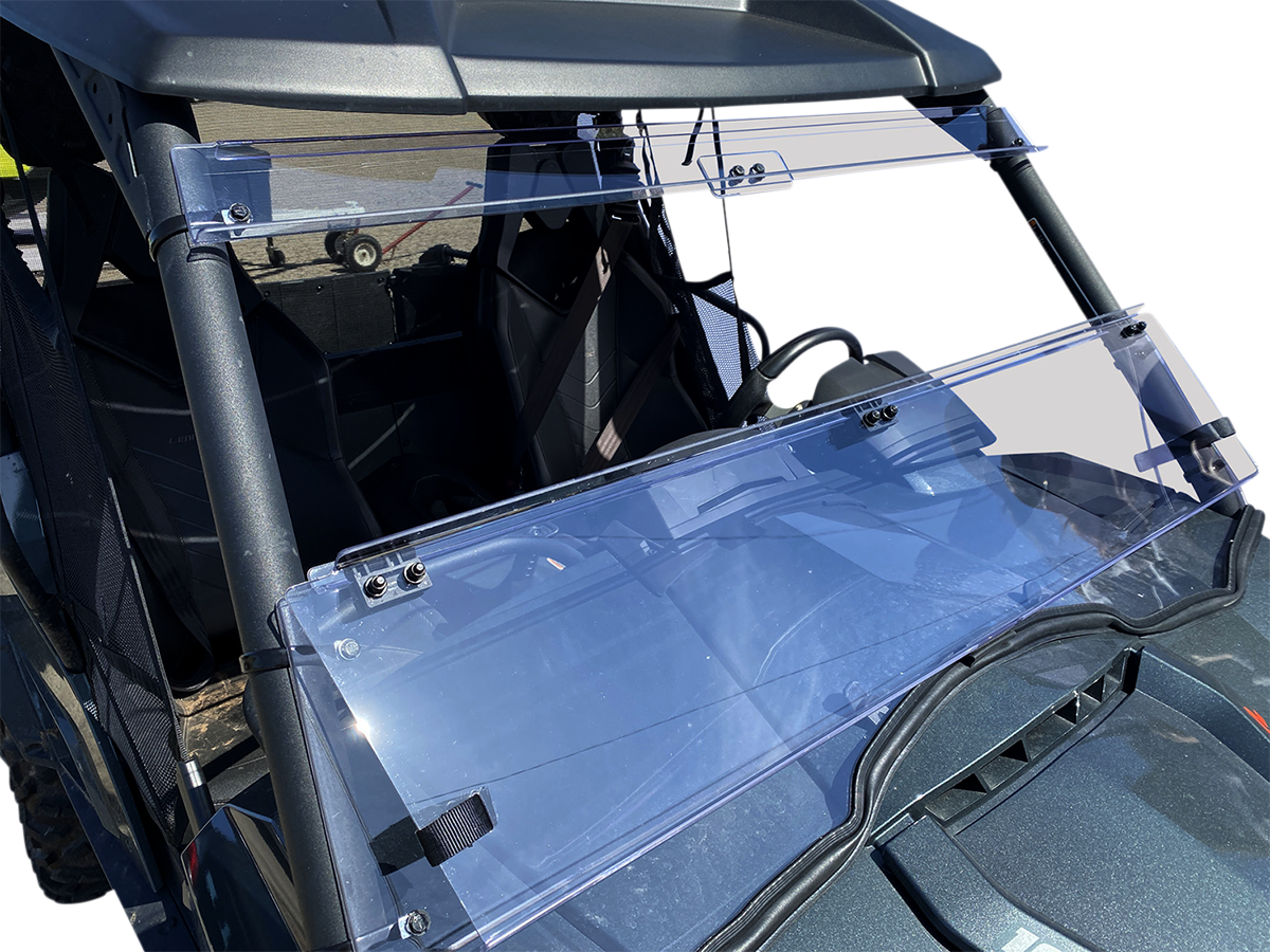 A close-up view of a utility vehicle’s clear polycarbonate folding windshield, mounted on a black roll cage. The windshield is partially open, showing two hinge sections and metal mounting brackets. The seats and steering wheel are visible behind the wind