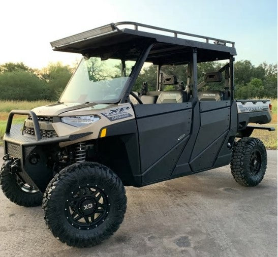 A Polaris Ranger Pro-Fit Full Size Crew with a beige and black metal top, four doors, and rugged tires is parked outdoors on pavement, surrounded by trees and grass under a partly cloudy sky.