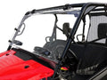 A close-up of a red Honda Pioneer 1000 with a Dual Vent Hard Coat Scratch Resistant Windshield (2015+), clear and fully attached to the black steel frame using multiple clamps. Black seats, mesh side nets, steering wheel, and dashboard controls are visibl