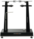 A black metal truing stand with adjustable uprights and pointed support pins holds a bicycle wheel hub axle horizontally. A metal gauge arm extends from the left side. The base is labeled “Moose Racing” and features bolts and a knurled knob for adjustment