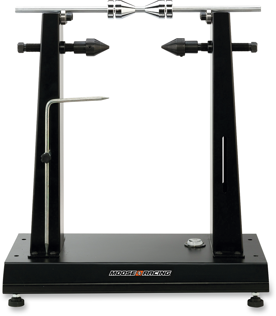 A black metal truing stand with adjustable uprights and pointed support pins holds a bicycle wheel hub axle horizontally. A metal gauge arm extends from the left side. The base is labeled “Moose Racing” and features bolts and a knurled knob for adjustment