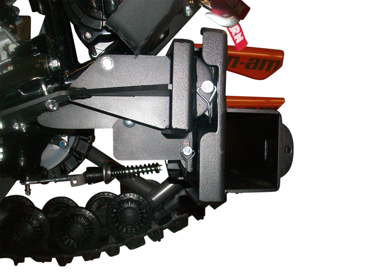 Close-up view of a mechanical component with a black metal bracket, bolts, spring, and part of a rubber track system. An orange part labeled Can-am is partially visible in the background, with a white background behind the machinery.