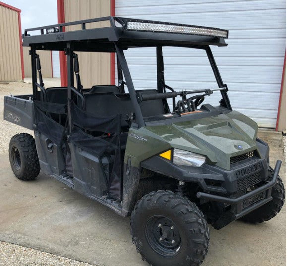 A Polaris Ranger Crew with a Mid-Size Crew Metal Roof (Pro-Fit Frame), olive green body, four seats, black mesh doors, rugged tires, and LED light bar is parked on concrete near a beige metal building and white garage door.