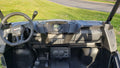 Close-up of the Arctic Cat Prowler Pro (2018-2022) with Defrost - Ice Crusher Cab Heater: dashboard features digital display, cup holders, control switches. Green grass and an asphalt road are visible in the sunlit background.