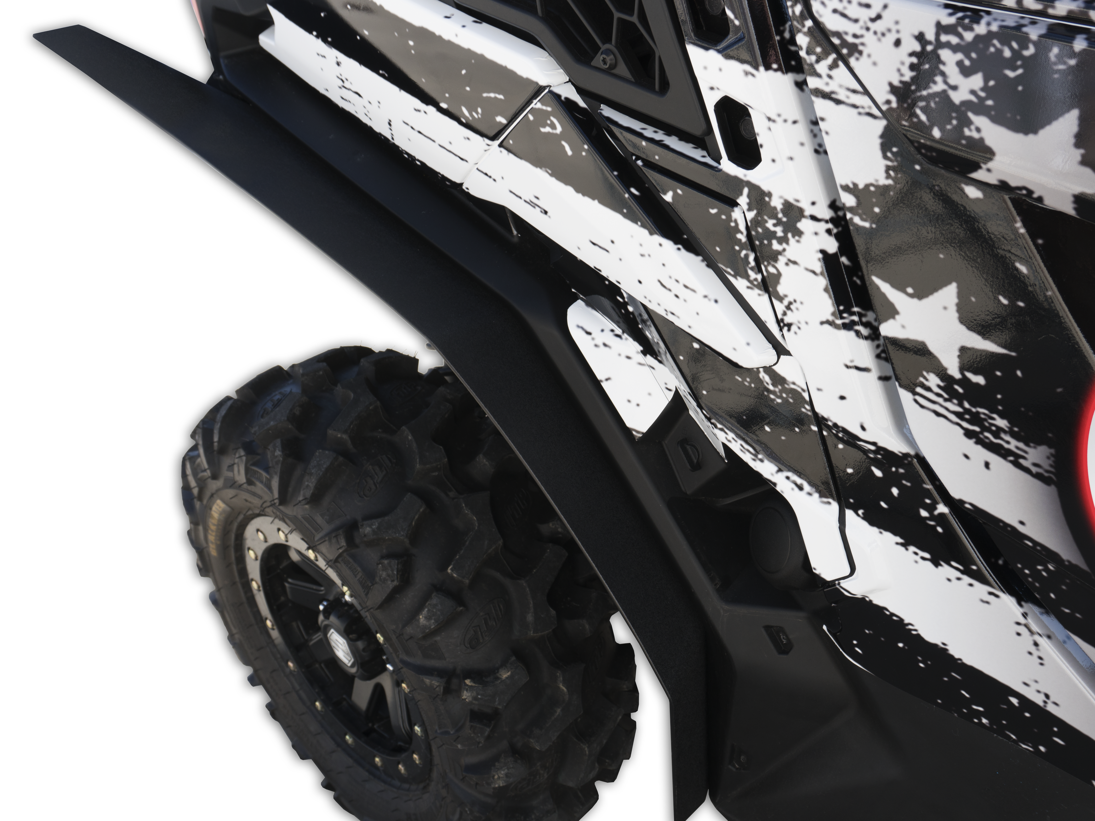 Close-up of Can Am Maverick Trail Over Fenders (Set 4) 2018+ on a rugged off-road vehicle. The large, knobby tire is built for rough terrain. The black and white splattered camo body features white stars and a partial red tail light.