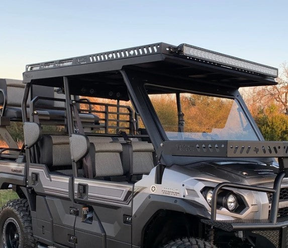 The Kawasaki Mule Pro Fxt Metal Roof 2015+ is a rugged, black and gray off-road utility vehicle with large tires, open cabin, reinforced gray-padded seats, heavy-duty roof rack with LED light bar, and a sunset sky with trees in the background.