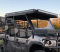 The Kawasaki Mule Pro Fxt Metal Roof 2015+ is a rugged, black and gray off-road utility vehicle with large tires, open cabin, reinforced gray-padded seats, heavy-duty roof rack with LED light bar, and a sunset sky with trees in the background.