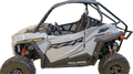 A silver Polaris RZR Trail S side-by-side off-road vehicle with black roll cage, rugged tires, and red suspension springs, viewed from the left side. The door features bold black RZR graphics and the word Polaris appears near the bottom.