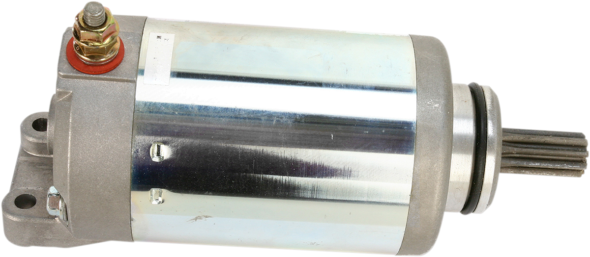 A metallic cylindrical starter motor with a shiny silver body, a terminal bolt with a red washer on top, mounting holes on the left end, and a protruding gear shaft on the right end, all set against a plain white background.