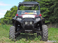 A rugged black Polaris RZR equipped with the Polaris RZR 800 to RZR S 800 5 Long Travel Kit is parked on grass, featuring large knobby tires and a roof. SuperATV logos are visible on the windshield and lower right. Trees and sky in background.