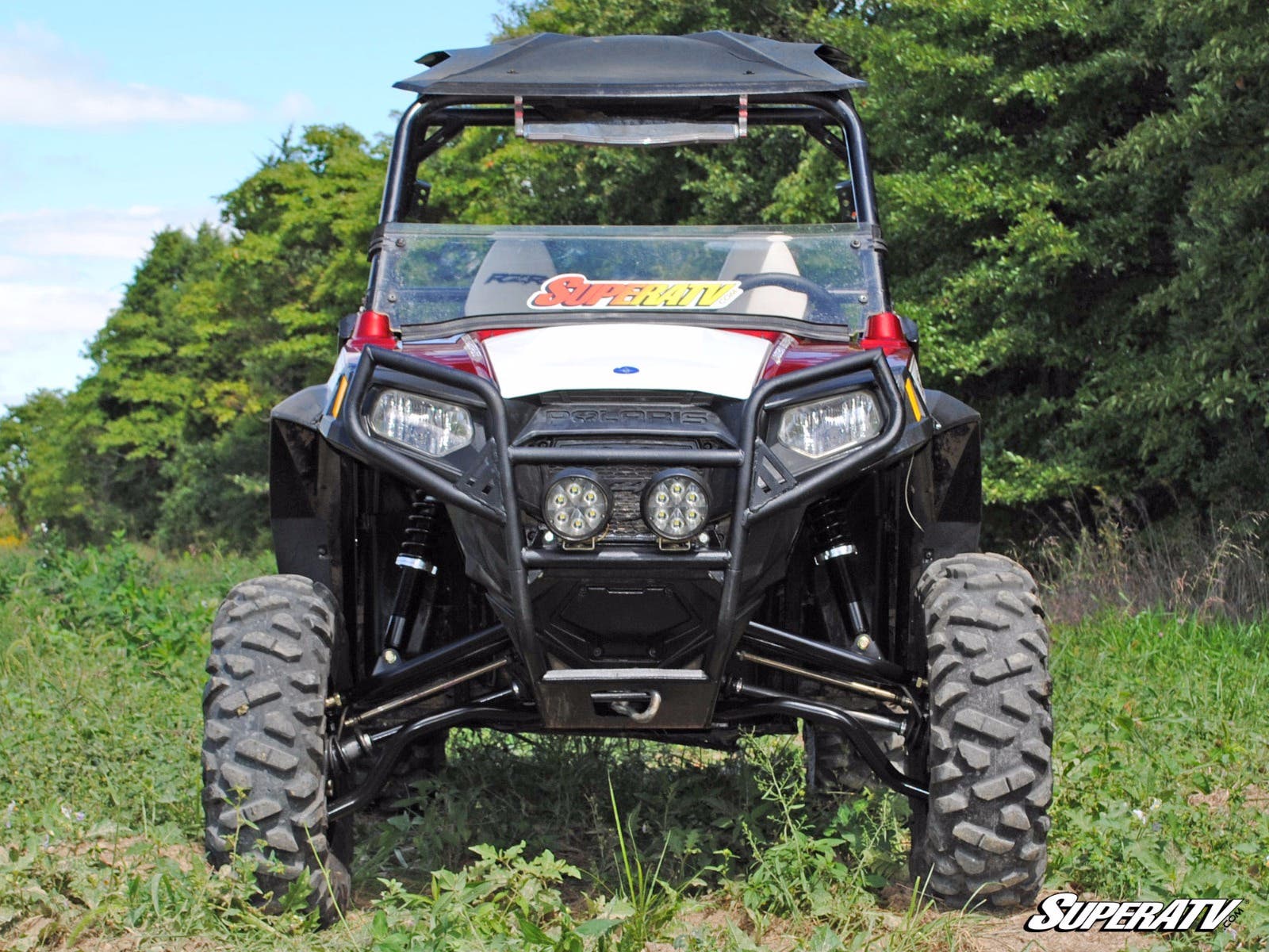 A rugged black Polaris RZR equipped with the Polaris RZR 800 to RZR S 800 5 Long Travel Kit is parked on grass, featuring large knobby tires and a roof. SuperATV logos are visible on the windshield and lower right. Trees and sky in background.