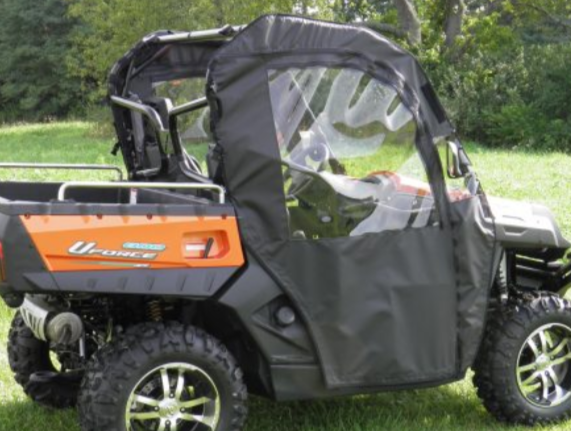 A CF Moto UForce 500/800 utility vehicle with large off-road tires is parked on grass, equipped with black soft doors and a clear plastic window. Black and orange in color, it sits amid trees and greenery in the background.