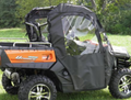 A CF Moto UForce 500/800 utility vehicle with large off-road tires is parked on grass, equipped with black soft doors and a clear plastic window. Black and orange in color, it sits amid trees and greenery in the background.