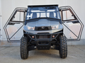 The Kymco UXV 700i Sport Cab Kit, a rugged grey off-road utility vehicle with large treaded tires and upward-opening glass doors, is parked on concrete in front of a plain white wall with a low metal ledge.