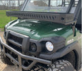 A green Kawasaki ATV with rugged tires is parked outdoors, featuring a sturdy black grill guard and a Kawasaki Mule Pro Front Roll Bar Basket mounted above the hood, with a mesh windshield behind it. Grass, gravel, and houses appear in the background.