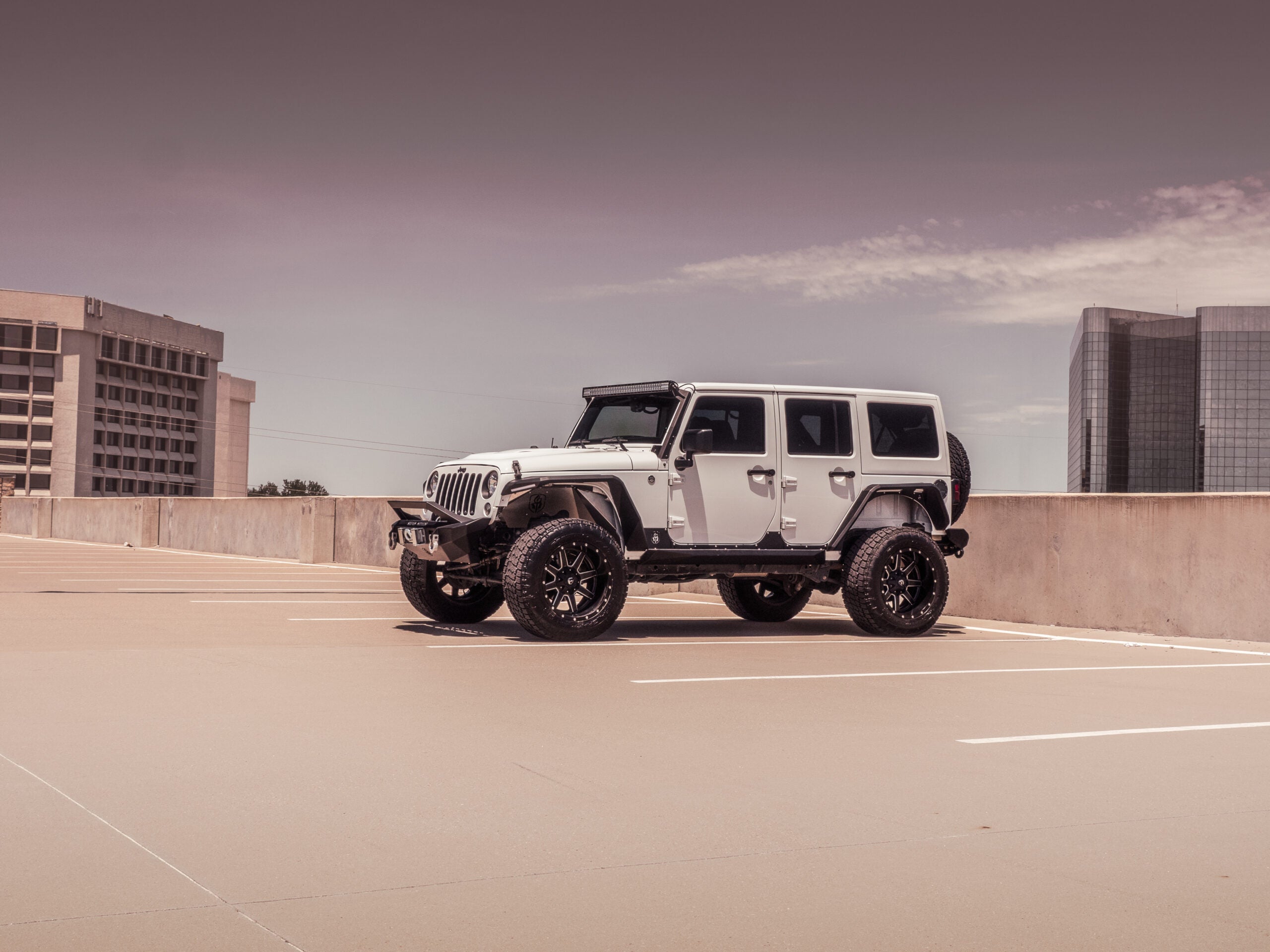 A white Jeep Wrangler with a 2007–2018 JK Stealth Front Winch Bumper Sheetmetal Bar Guard in texture black and large off-road tires is parked atop a nearly empty garage, with modern offices in the background beneath a partly cloudy sky.