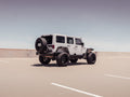 A white, four-door Jeep Wrangler with a textured black 2007-2018 Jeep JK Stealth Rear Bumper Mid Width with Tire Carrier Assembly is parked alone in a sunlit rooftop lot, showcasing its oversized tires and lifted suspension.