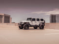 A 2007-2018 Jeep JK with Stealth Front Fender Flare Body Armor and Switchback LED DRL (Texture Black) is parked in an empty rooftop lot under an overcast sky, modern office buildings in the background, centered and facing left.
