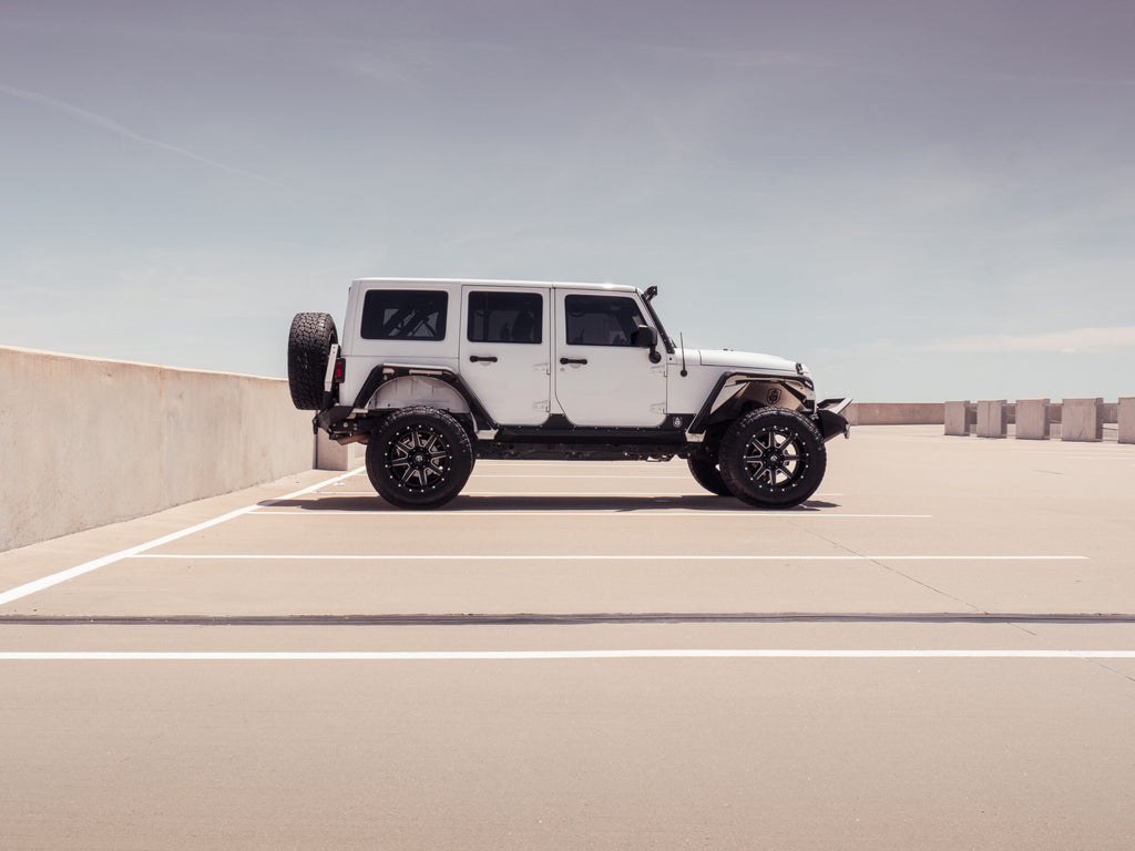 A 2007-2018 Jeep JKU 4-door, equipped with Stealth Rear Fender Flare Body Armor (Texture Black), is parked in an open-air lot under a clear sky, centered in the frame. The bright scene highlights the Jeeps rugged black accents.