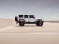 A 2007-2018 Jeep JKU 4-door, equipped with Stealth Rear Fender Flare Body Armor (Texture Black), is parked in an open-air lot under a clear sky, centered in the frame. The bright scene highlights the Jeeps rugged black accents.