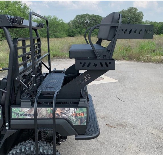 A Kawasaki Mule Pro FXT with a High Seat is parked on pavement. The custom elevated metal seat with armrests and a footrest is mounted rear-facing, overlooking a grassy field, trees, and cloudy sky in the background.