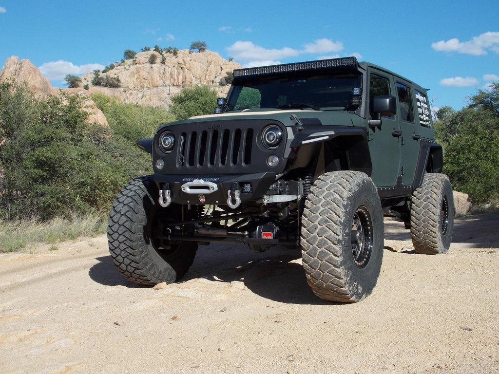 A matte green Jeep Wrangler with a lifted suspension, oversized off-road tires, and a 2018+ Stealth Front Winch Bumper (Texture Black Warn Vr Evo) is parked on a dirt trail among shrubs and rocky hills under a clear blue sky.