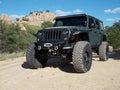 A matte green Jeep Wrangler with a lifted suspension, oversized off-road tires, and a 2018+ Stealth Front Winch Bumper (Texture Black Warn Vr Evo) is parked on a dirt trail among shrubs and rocky hills under a clear blue sky.
