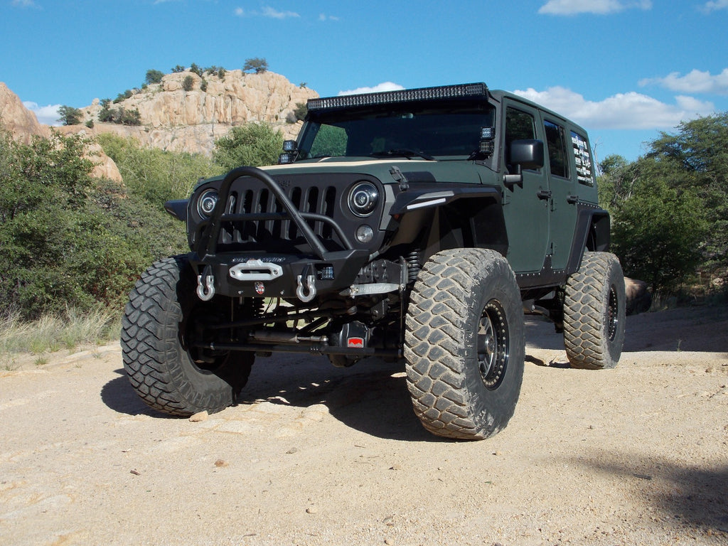 A matte green Jeep Wrangler with large off-road tires, roof LED light bar, and a 2007+ JL Rubicon Stealth Front Winch Bumper Stinger Guard Competition Cut (Texture Black Warn Vr Evo) is parked on a rocky desert trail under a blue, partly cloudy sky.