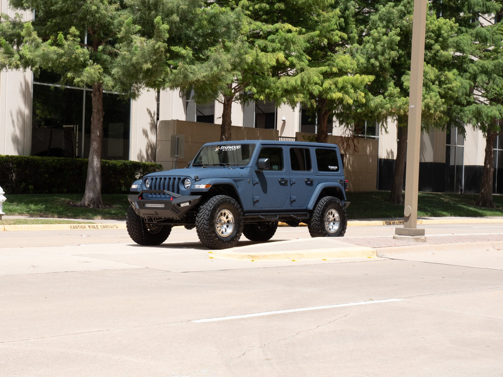 A blue Jeep Wrangler with off-road tires and a 2018+ Jeep JL Sport Spartan Front Bumper in texture black is parked by the curb on a sunny street, with green trees and a beige office building with reflective windows in the background.