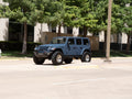 A blue Jeep Wrangler with off-road tires and a 2018+ Jeep JL Sport Spartan Front Bumper in texture black is parked by the curb on a sunny street, with green trees and a beige office building with reflective windows in the background.