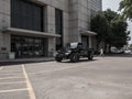 A 2018+ Jeep JL Sport with a Road Armor Spartan front bumper and pre-runner guard in texture black is parked outside a modern glass building, as a woman walks by and trees cast shade nearby in the bright urban scene.