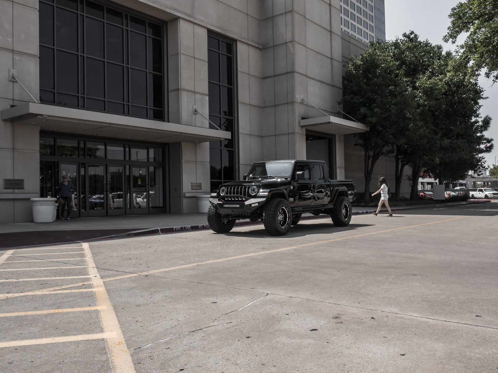 A 2018+ Jeep JL Sport with a Road Armor Spartan front bumper and pre-runner guard in texture black is parked outside a modern glass building, as a woman walks by and trees cast shade nearby in the bright urban scene.
