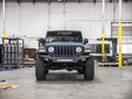 A matte black Jeep Wrangler with large off-road tires and a 2018+ Jeep JL Sport Spartan Front Bumper Bolt-On Accessory Skid Plate Guard by Road Armor is parked in a spacious industrial garage with scattered equipment, yellow pillars, and concrete floors.