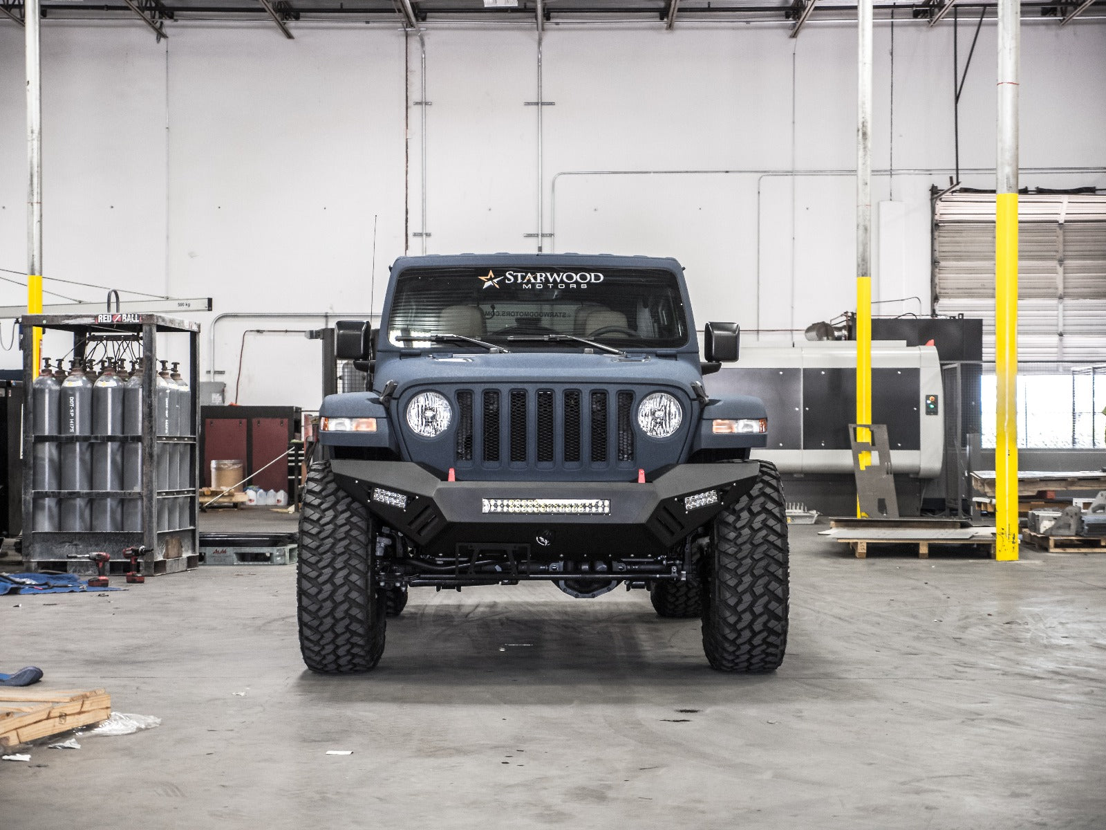 A matte black Jeep Wrangler with large off-road tires and a 2018+ Jeep JL Sport Spartan Front Bumper Bolt-On Accessory Skid Plate Guard by Road Armor is parked in a spacious industrial garage with scattered equipment, yellow pillars, and concrete floors.