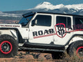 A white 2018+ Jeep JL Rubicon with Stealth JL Rocker Armor Panel and Guard (4 Door, Texture Black) is parked on rocky terrain outdoors. Snow-capped mountains and a clear blue sky are visible in the background, showing its front and side.