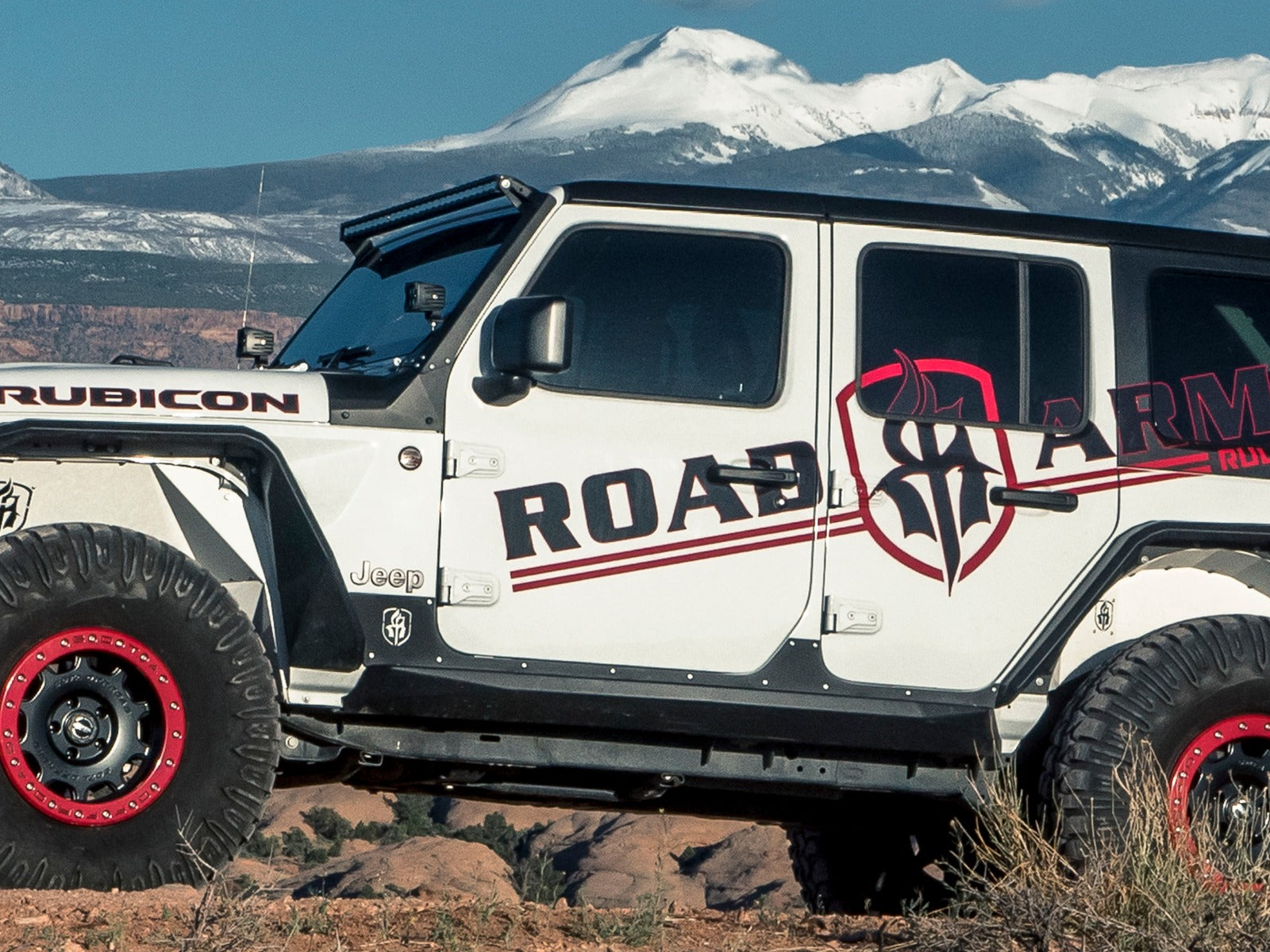 A white 2018+ Jeep JL Rubicon with Stealth JL Rocker Armor Panel and Guard (4 Door, Texture Black) is parked on rocky terrain outdoors. Snow-capped mountains and a clear blue sky are visible in the background, showing its front and side.