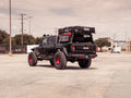 A black Jeep JT Sport with a Stealth Rear Winch Bumper and 9500lb remote winch is parked in an empty lot, featuring red wheel rims, off-road tires, roof tent attachment, and camping gear in the bed. Power lines and trees are in the background.