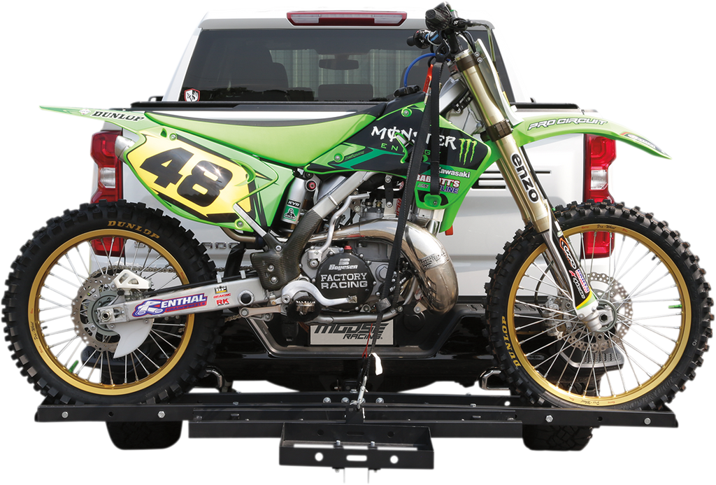 A green motocross bike with number 48, featuring Monster Energy branding, is securely mounted on a hitch carrier attached to the back of a white pickup truck. The bike’s tires and details are clearly visible against the clean truck background.