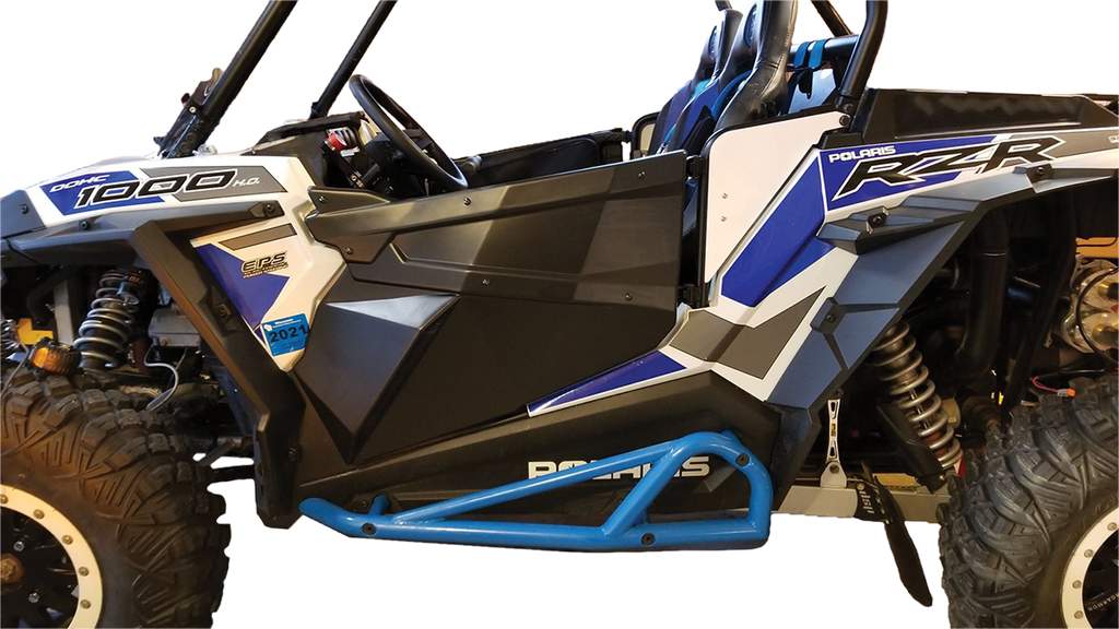 A close-up side view of a blue, white, and black Polaris RZR off-road vehicle, highlighting its rugged tires, suspension, roll cage, graphics, and blue rock slider, with the drivers seat and steering wheel partially visible.