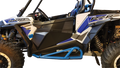 A close-up side view of a blue, white, and black Polaris RZR off-road vehicle, highlighting its rugged tires, suspension, roll cage, graphics, and blue rock slider, with the drivers seat and steering wheel partially visible.