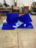 Four large, shiny, deep blue Rzr XP 4 1000 doors with angular shapes rest on a blue blanket on a workshop floor surrounded by industrial machines, workbenches, and tools in a spacious workshop with concrete flooring and white walls.
